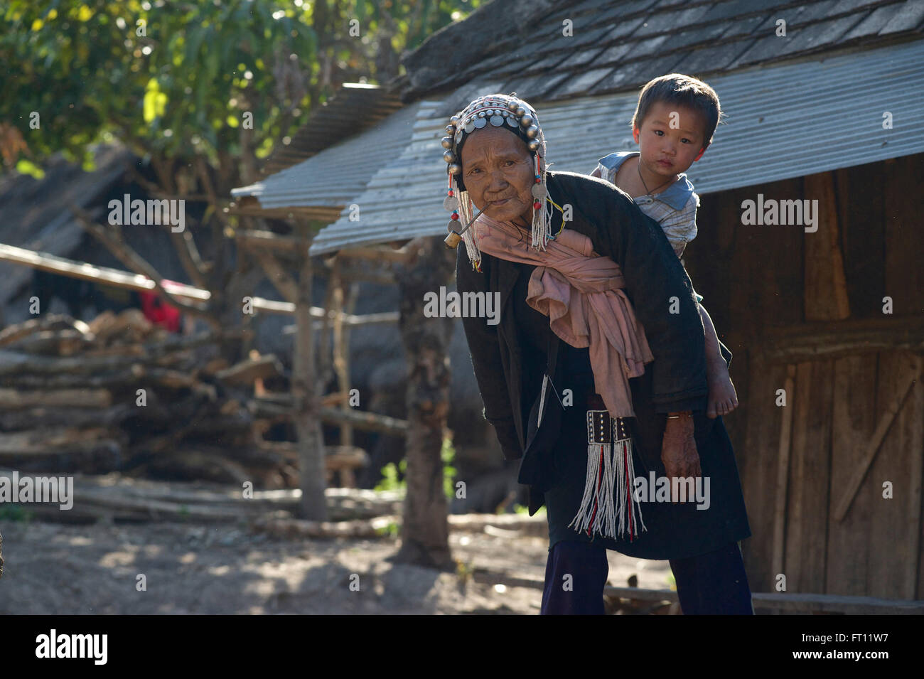 Alten Akha-Frau in traditioneller Kleidung zurück raucht eine Pfeife und tragen ihr Enkelkind auf ihr in der Nähe von Kyaing Tong, Kentung, Shan State in Myanmar Burma Stockfoto