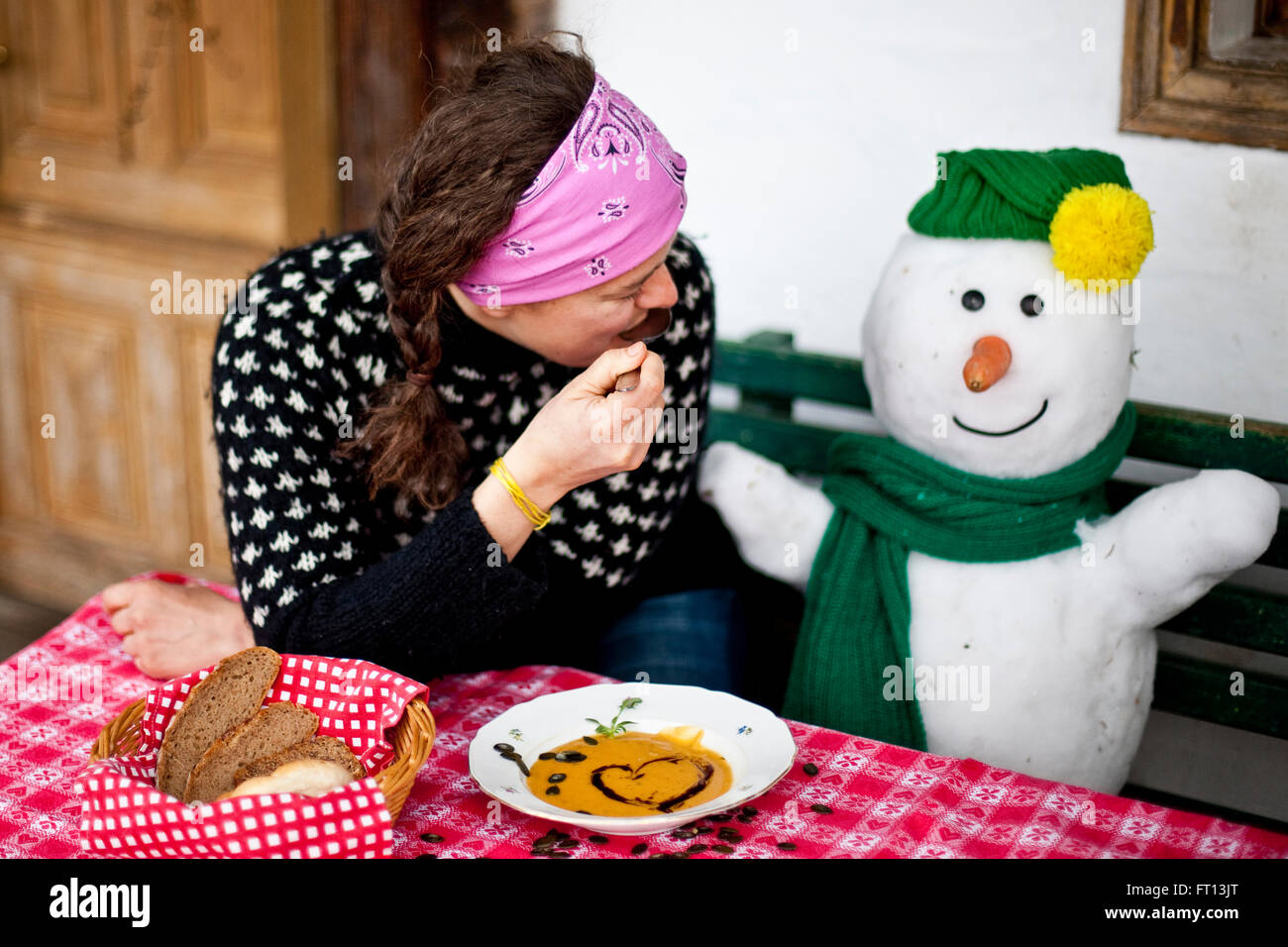 Frau sitzt auf einer Bank neben einem Schneemann während des Essens Kürbis Cremesuppe, Steiermark, Österreich Stockfoto