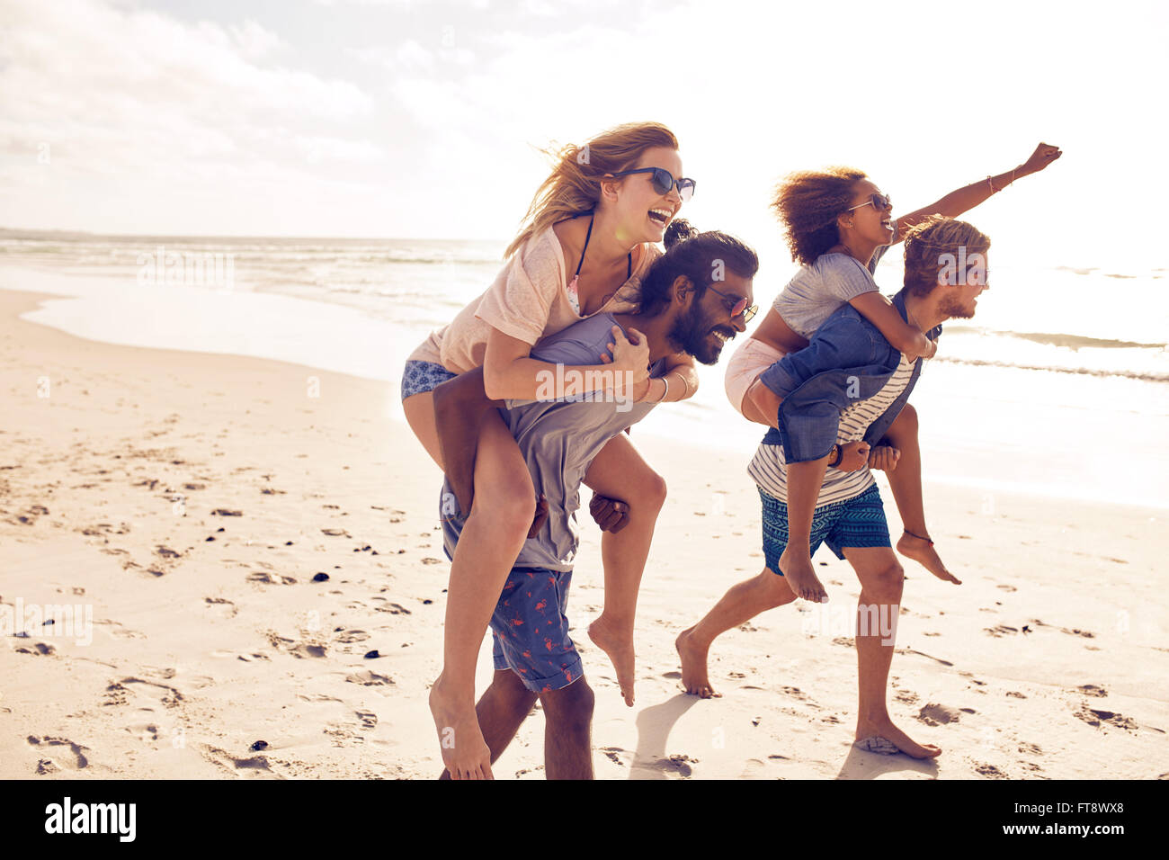 Zwei junge Männer geben ihre Freundinnen Huckepack tragen am Strand. Fröhlichen jungen Freunde im Sommer am Strand genießen. Stockfoto
