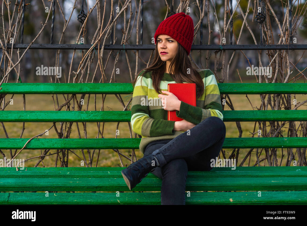 Teenager-Mädchen im roten Hut und grünen Pullover sitzen auf einer Bank in einem Park und hält ein rotes Buch. Mittlere Aufnahme. Stockfoto