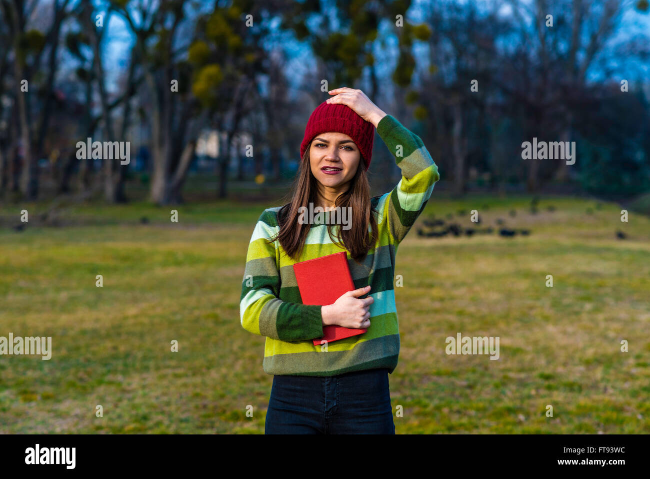 Ein Teenager-Mädchen im roten Hut und grün gestreifte Pullover Holding Buch mit rotem Einband stehen auf einer Wiese in einem Park. Stockfoto