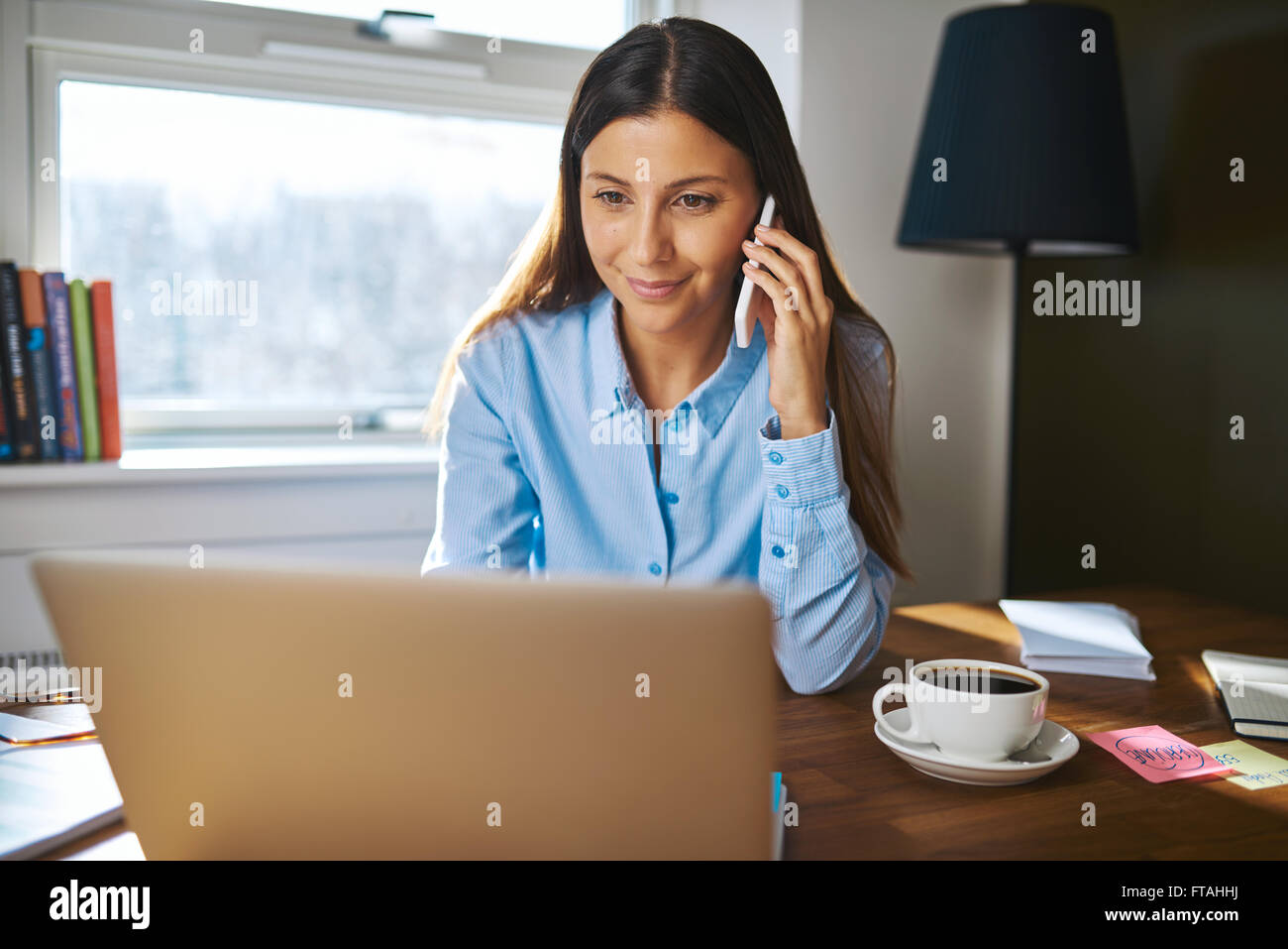 Fröhlicher junger weiblichen Erwachsenen Geschäft Inhaber mit Grinsen am Telefon während der Arbeit am Laptop Computer am Schreibtisch neben Kaffee Tasse mit w Stockfoto