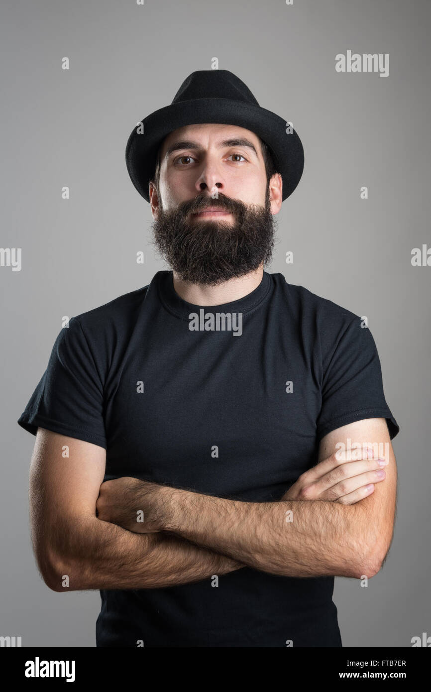 Harte bärtige Hipster mit verschränkten Armen trug schwarze T-shirt und Hut Blick in die Kamera. Kopfschuss-Porträt über grauen Hintergrund Stockfoto