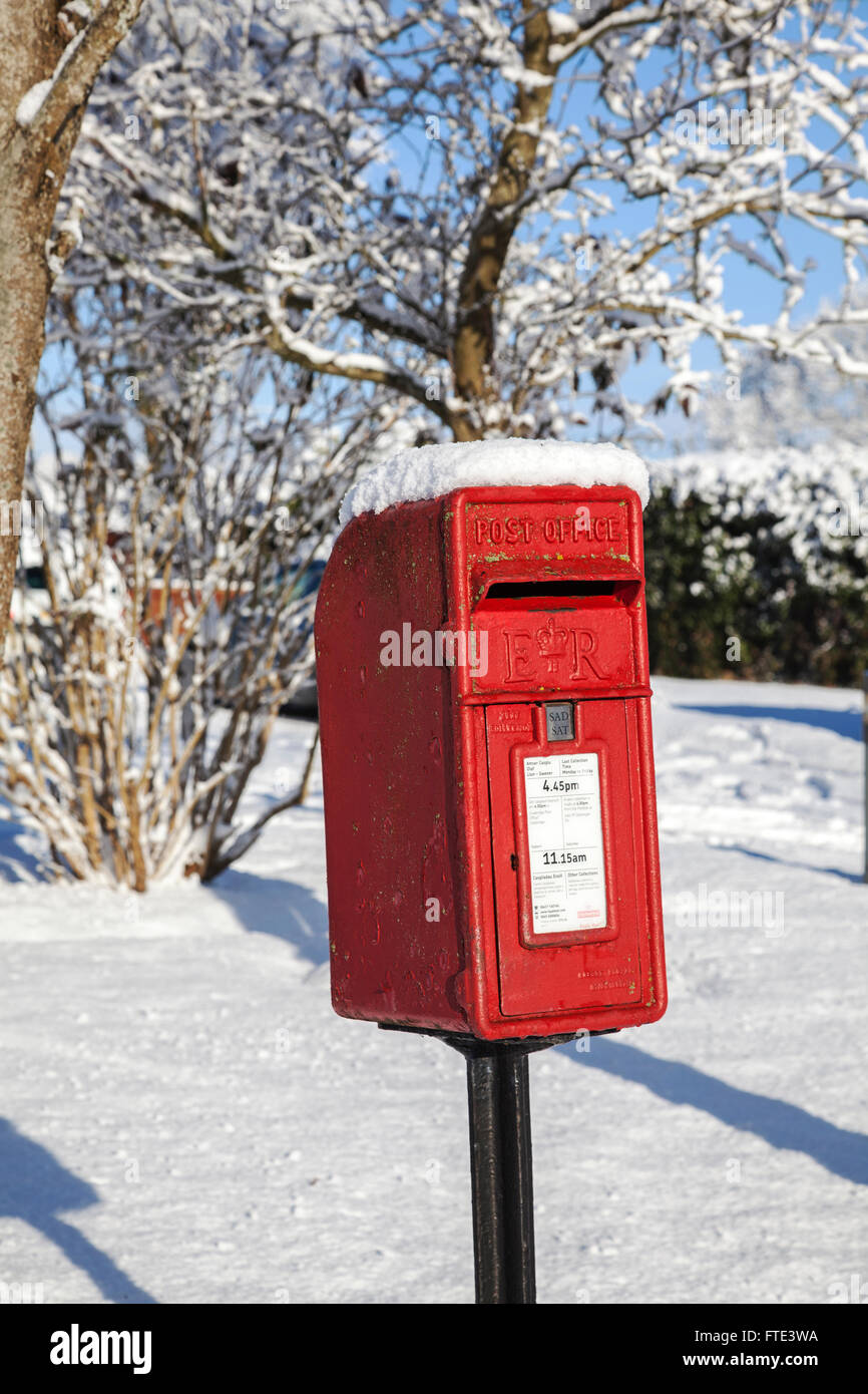 Roter Sockel Briefkasten in hellem Sonnenlicht in Schnee bedeckt Mit verdeckten Bäumen im Hintergrund Vor einem klaren blauen Himmel Stockfoto