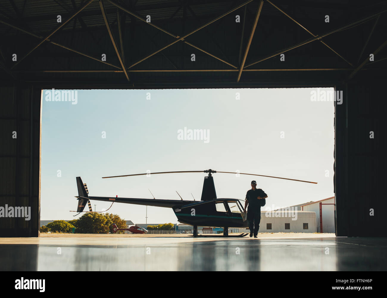 Silhouette eines Piloten im Hangar mit einem geparkten Hubschrauber laufen. Pilot, Ankunft am Flughafen mit einem Hubschrauber im Hintergrund. Stockfoto