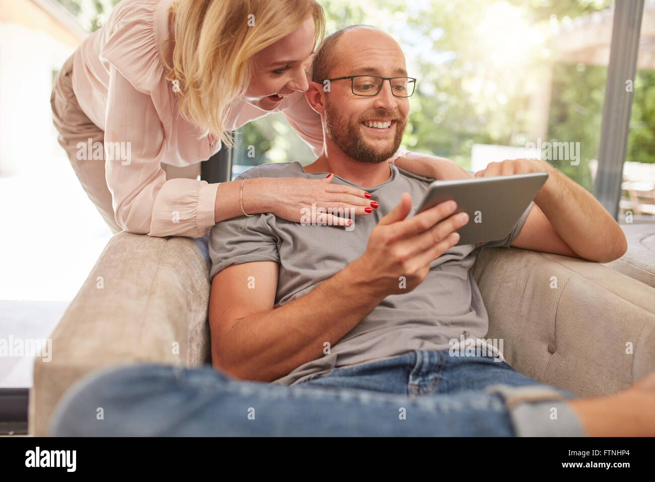 Glückliche Frau hinter ihrem Ehemann sitzt der Sofa im Wohnzimmer, beide auf der Suche auf eine digitale Tablet lächelnd. Stockfoto