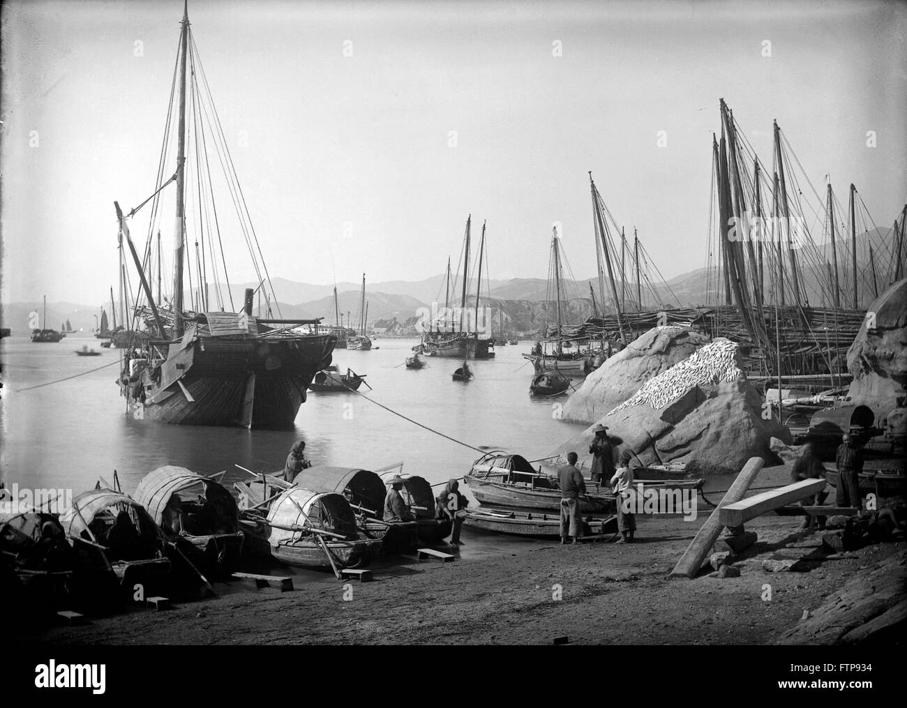 AJAXNETPHOTO. 1890 (CA.). HONG KONG. EINE SZENE MIT EINER GROßEN HANDEL DSCHUNKE VOR ANKER STERN-AN DER KÜSTE, MIT KLEINEREN FISCHEN SAMPANS AM STRAND AUFGESTELLT. AUF DER RECHTEN SEITE AUF EINEM FELSEN TROCKNET EIN FANG VON FISCHEN IN DER SONNE, WÄHREND EIN TISCHLER SCHIFFBAUER GERADE FERTIG SCHNEIDEN EINE GROßE HOLZ, MÖGLICHERWEISE FÜR JUNK-E-GEBÄUDE IN DER NÄHE.   FOTO: AJAX VINTAGE BILD BIBLIOTHEK REF: JUNKS 01 Stockfoto
