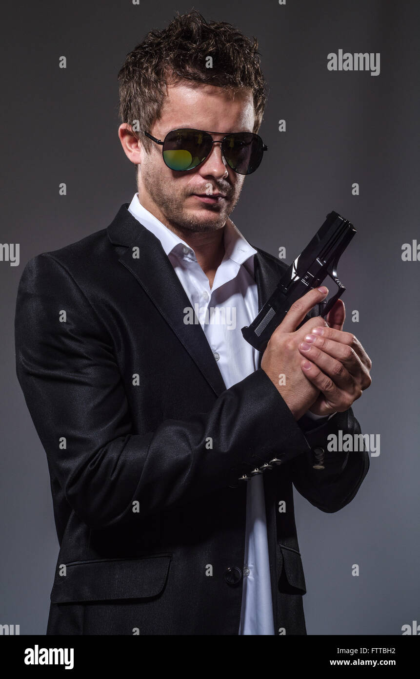 Mann im dunklen Anzug mit Pistole und dunklen reflektierenden Sonnenbrille auf einfachen grauen Hintergrund halblängen erschossen Stockfoto