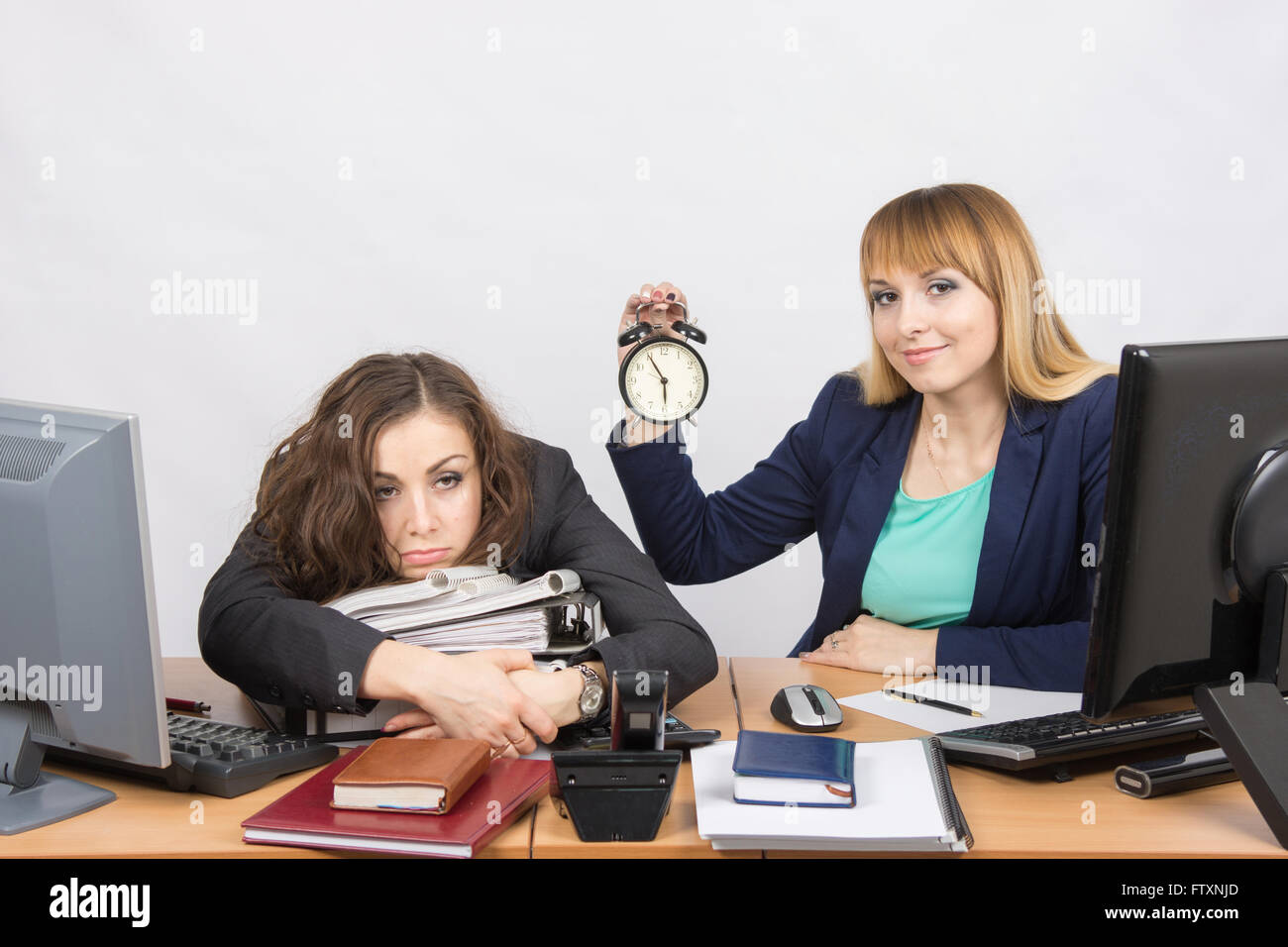 Zwei Mädchen im Büro am Ende des Tages, mit einem Lächeln, halten Sie eine Uhr, liegt ein anderes müde auf Ordner Stockfoto