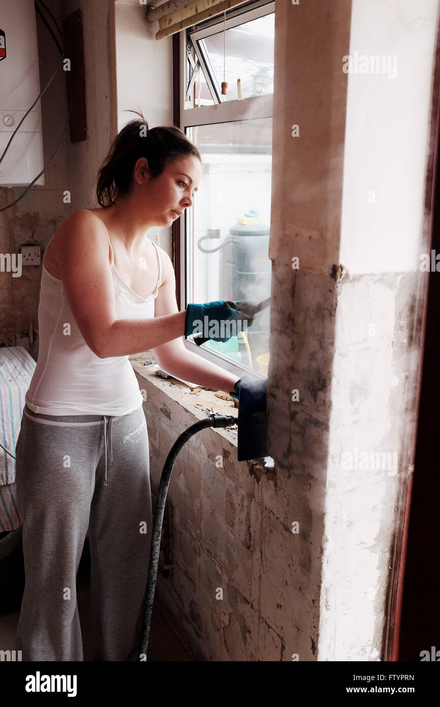 Junge Frau in ihren 20ern tun DIY in ihrem Bungalow Haus mit Dampfer und Schaber entfernen Wandfliesen in Küche Stockfoto