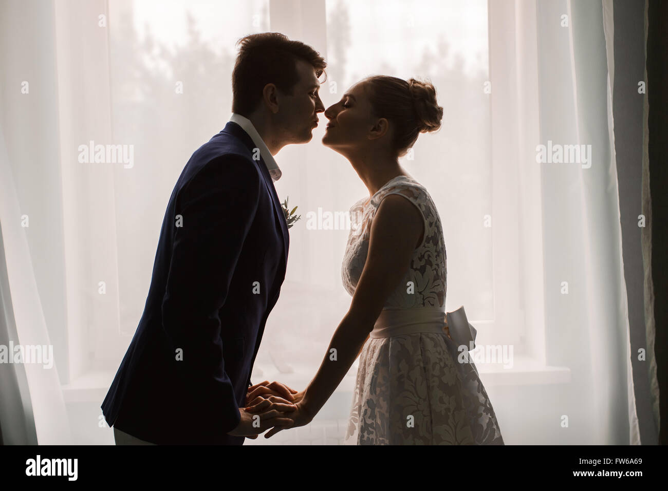 Glücklich Frischvermählten küssen indoor. Brünette Braut und Bräutigam stehen in der Nähe von Fenster-Silhouetten von glücklich Stockfoto