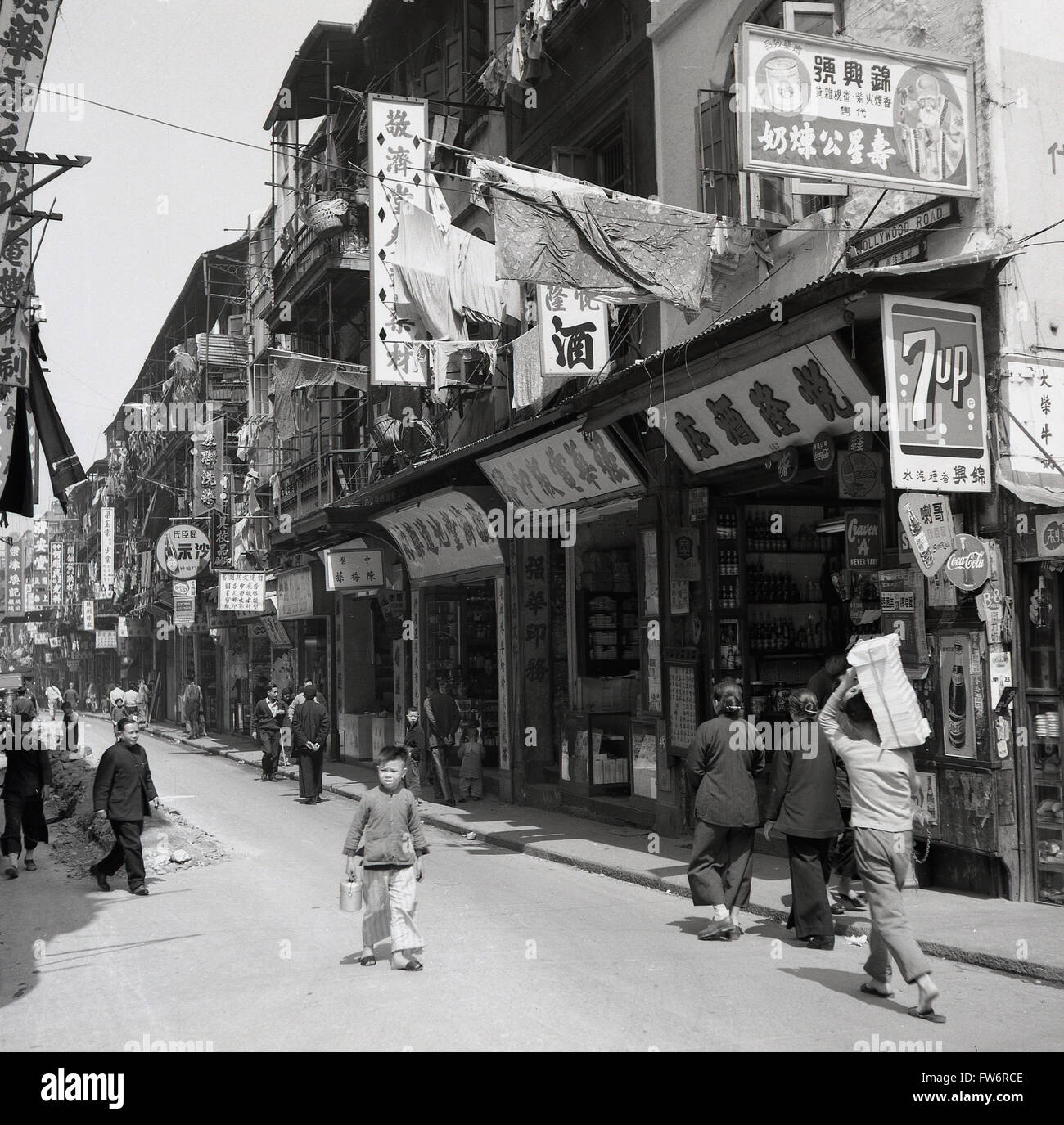 1950er Jahre, historische Bild, eine Straßenszene in Hongkong, die Geschäfte von der Zeit, die lebendige Unterkunft über Ihnen, Menschen vor Ort gehen und die vielen Werbe- und Hinweisschildern. Stockfoto