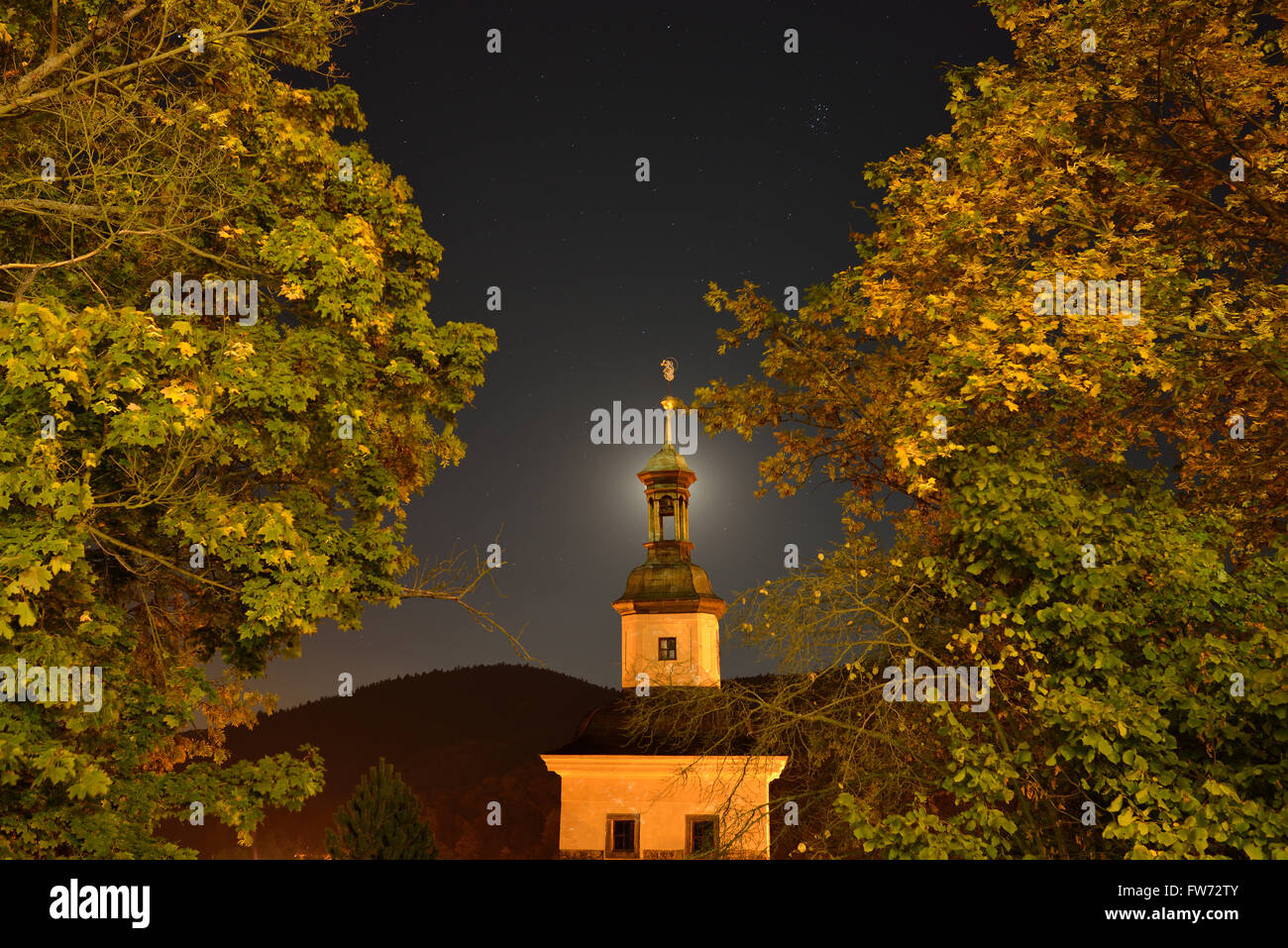 Kirche und Laub von Straßenlaternen mit dem Mond hinter dem Kirchturm beleuchtet. Loket, Bezirk Sokolov, Böhmen, Tschechische Republik. Stockfoto