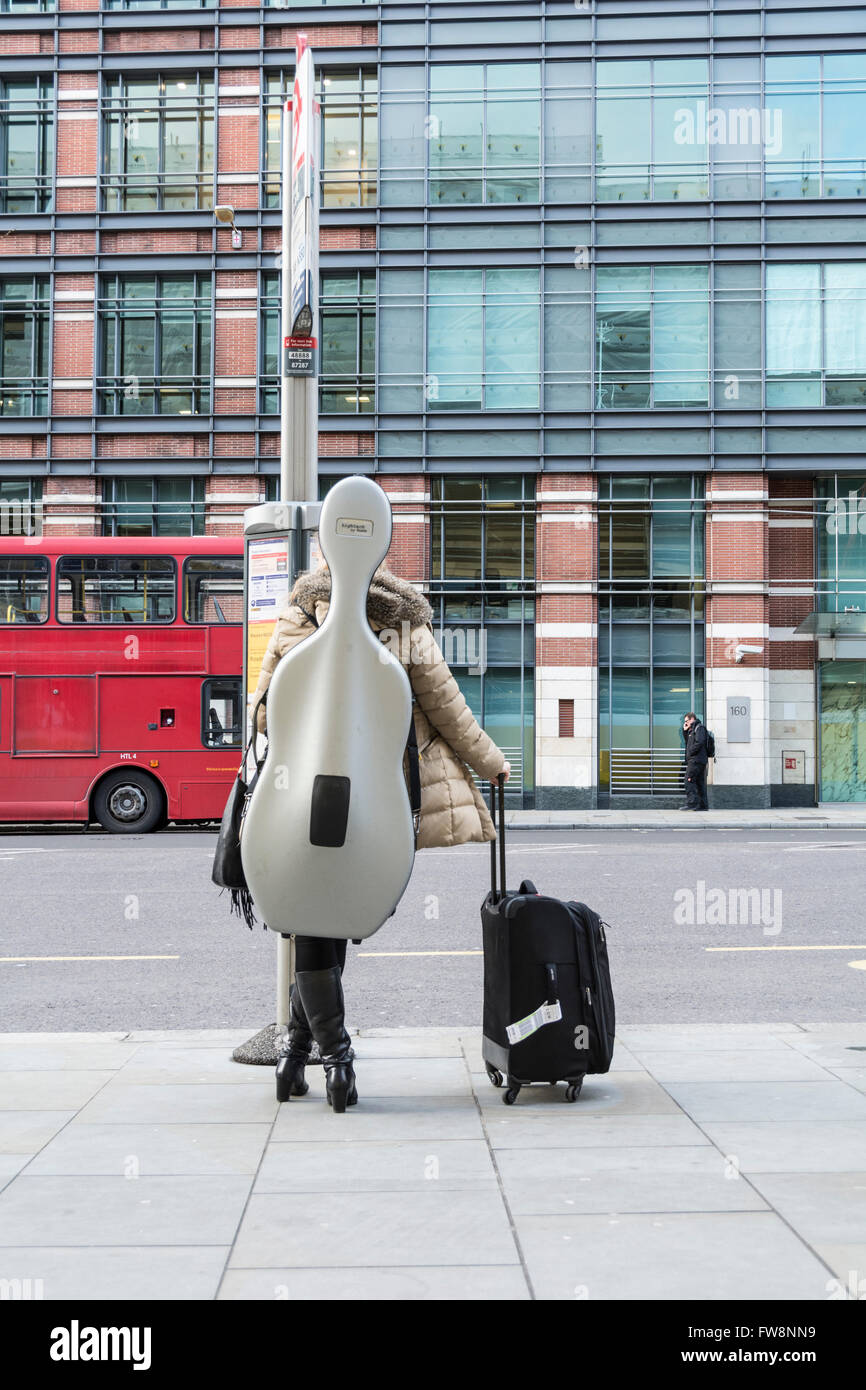 Cello Willen Reisen - eine junge Frau wartet auf ein Bus im Zentrum von London mit einem großen Cello an ihrem Rücken geschnallt. Stockfoto
