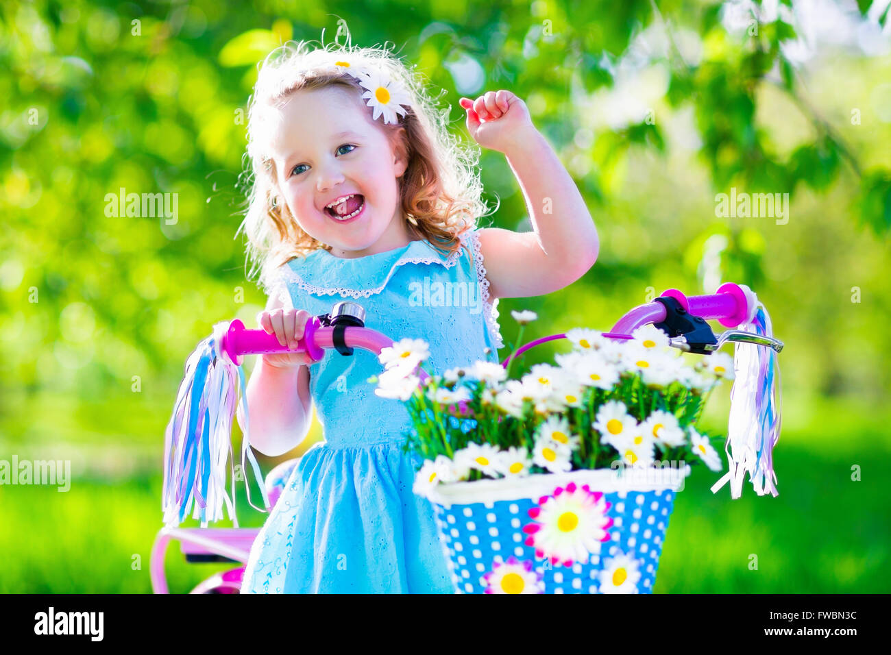 Glückliches Kind mit dem Fahrrad. Süßes Kind Radfahren im Freien. Kleines Mädchen in einem blauen Kleid auf einem rosa Fahrrad mit Daisy Blumen im Korb Stockfoto
