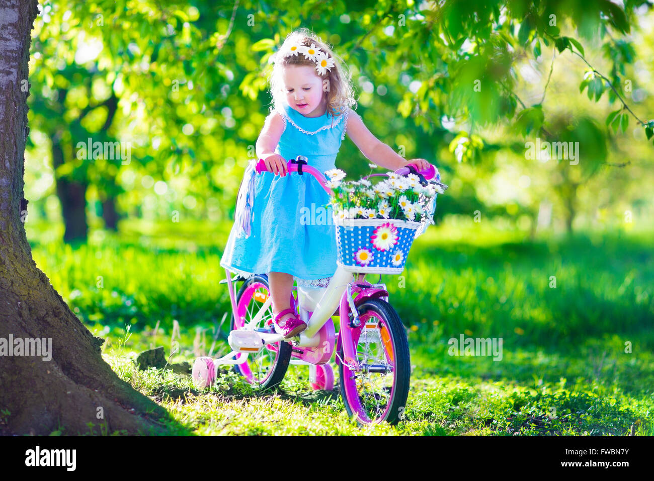 Glückliches Kind mit dem Fahrrad. Süßes Kind Radfahren im Freien. Kleines Mädchen in einem blauen Kleid auf einem rosa Fahrrad mit Daisy Blumen im Korb Stockfoto