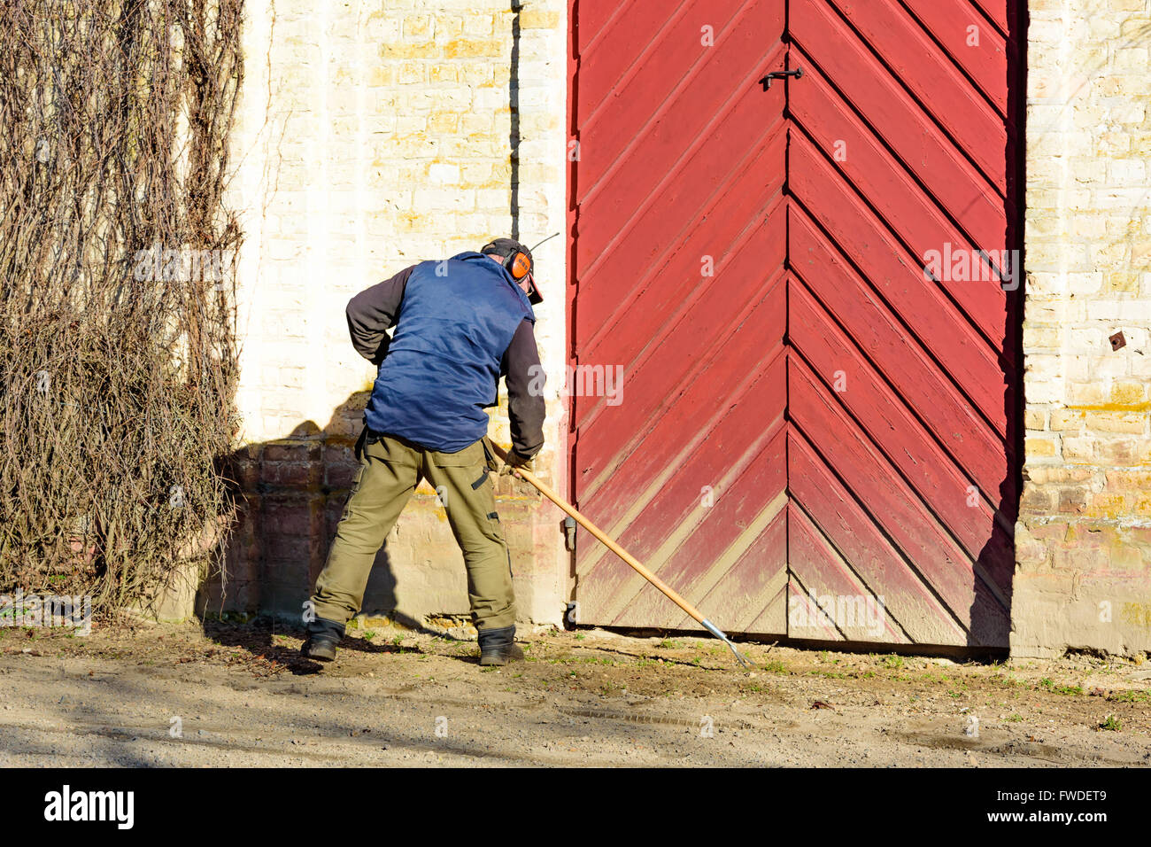 Vittskovle, Schweden - 1. April 2016: Mann ist eine Handgemenge Hacke außerhalb eines Gebäudes mit einem großen roten Tür verwenden. Er ist das Unkraut her entfernen Stockfoto