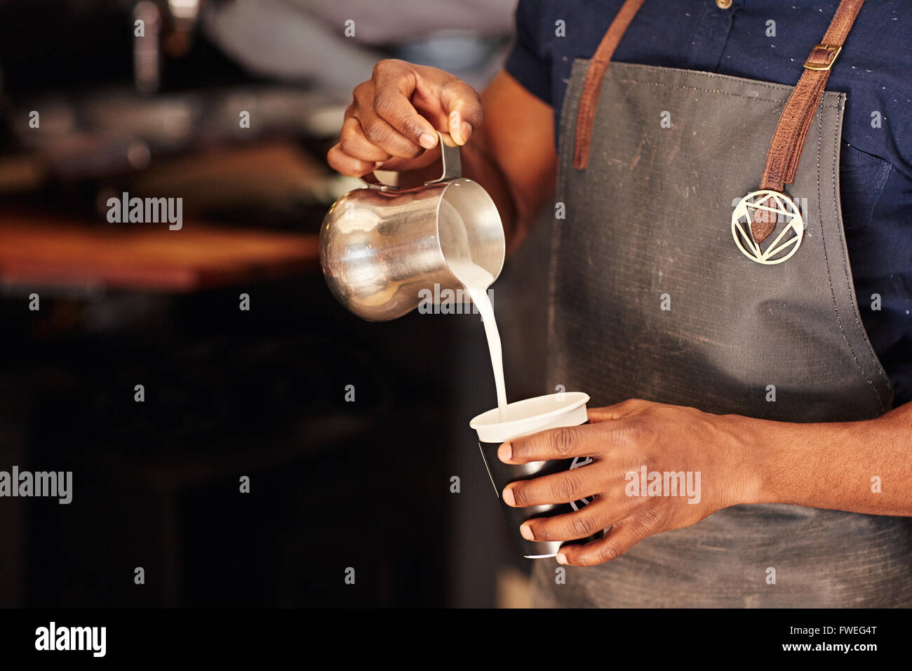 Bild von einem afrikanischen Barista sorgfältig gießt Milch aus einem Edelstahl-Krug in eine Take-away Tasse in einem Café beschnitten Stockfoto