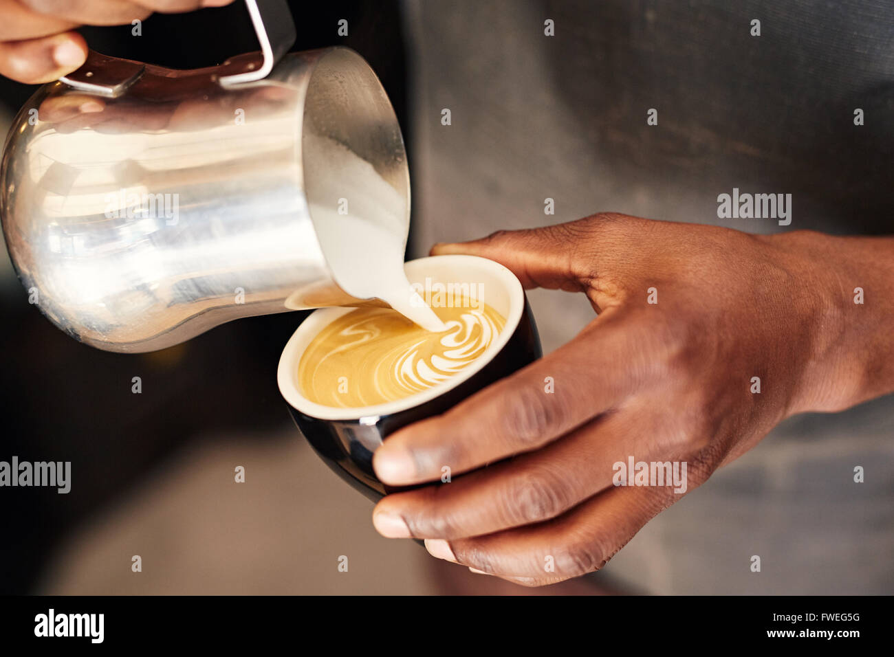 Hände von einer qualifizierten afrikanischen Barista strömenden Milchschaum mit einer Edelstahl-Kanne in einer Keramik Kaffeetasse und bilden eine attr Stockfoto