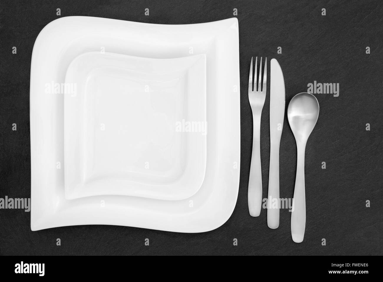 Moderne Tischdekoration mit Porzellanteller und Edelstahl-Besteck über Schiefer Hintergrund. Stockfoto