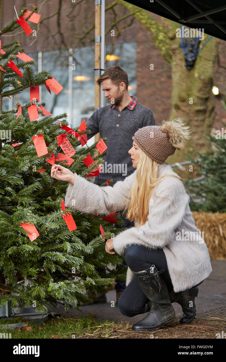 Eine Frau die Wahl einer traditionellen Kiefer, Blick auf die roten handgeschriebenen Etiketten auf einer Tanne Weihnachtsbaum. Stockfoto