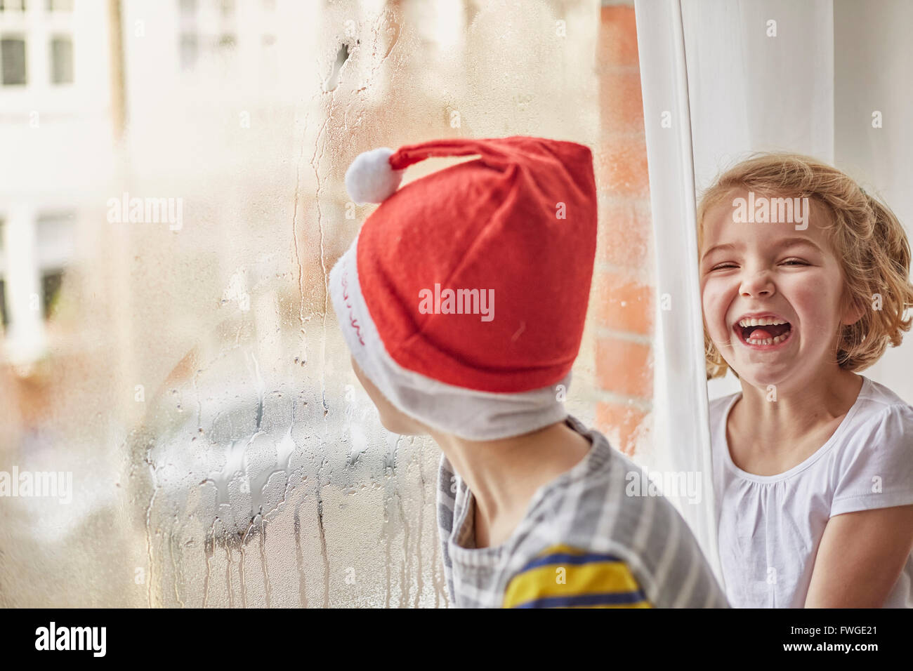 Weihnachtsmorgen in einem Einfamilienhaus. Ein Junge in eine Weihnachtsmütze aus einem Schlafzimmerfenster und seine Schwester neben ihm lachend. Stockfoto