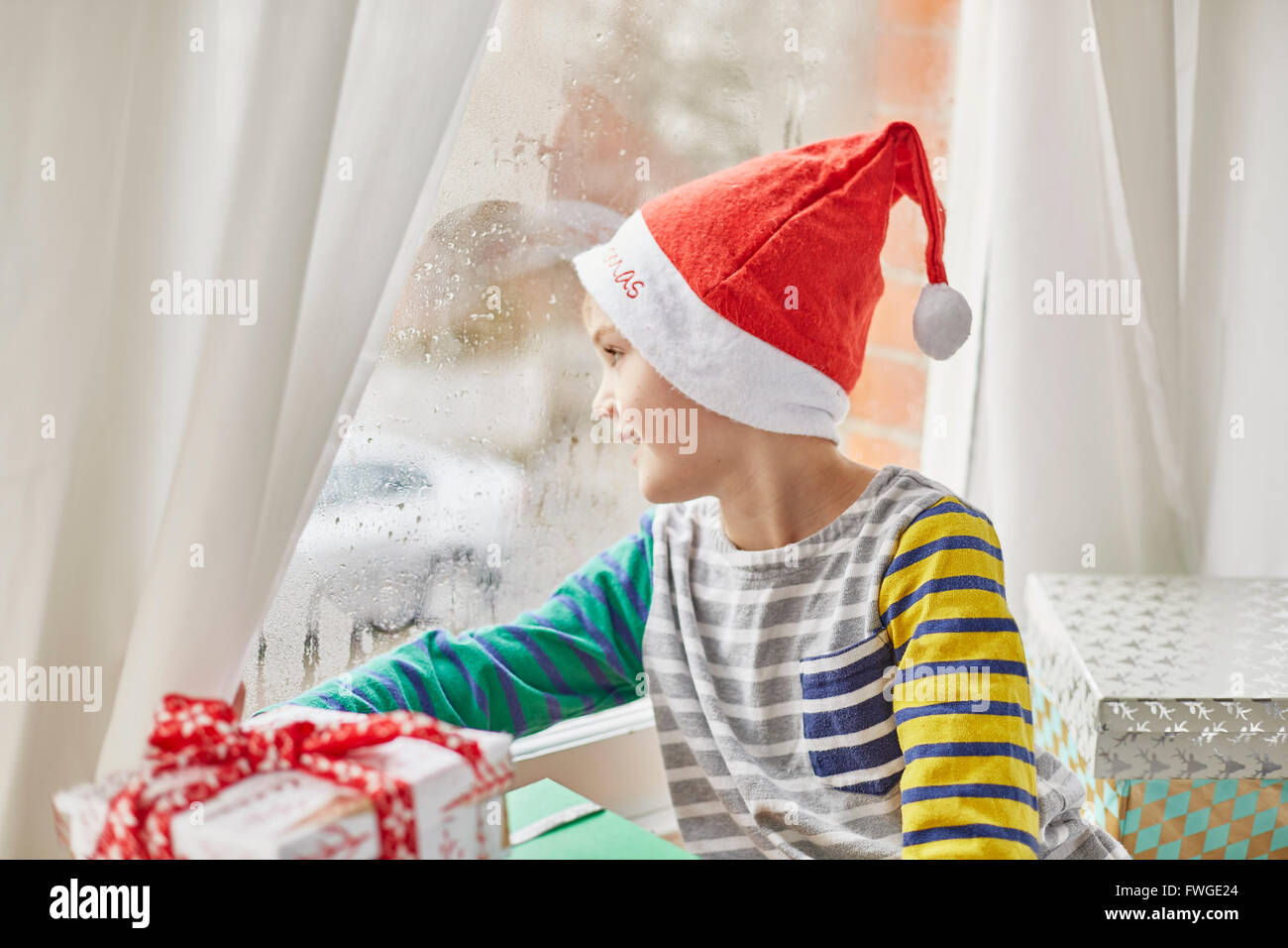 Weihnachtsmorgen in einem Einfamilienhaus. Ein Junge in eine Weihnachtsmütze Blick aus dem Schlafzimmerfenster. Stockfoto