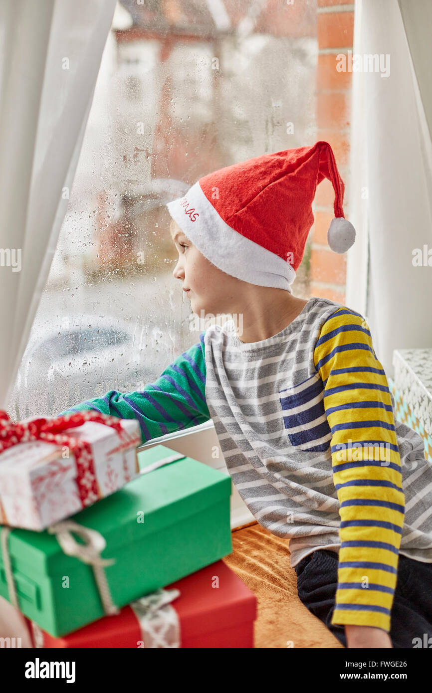 Weihnachtsmorgen in einem Einfamilienhaus. Ein Junge in eine Weihnachtsmütze Blick aus dem Schlafzimmerfenster. Stockfoto