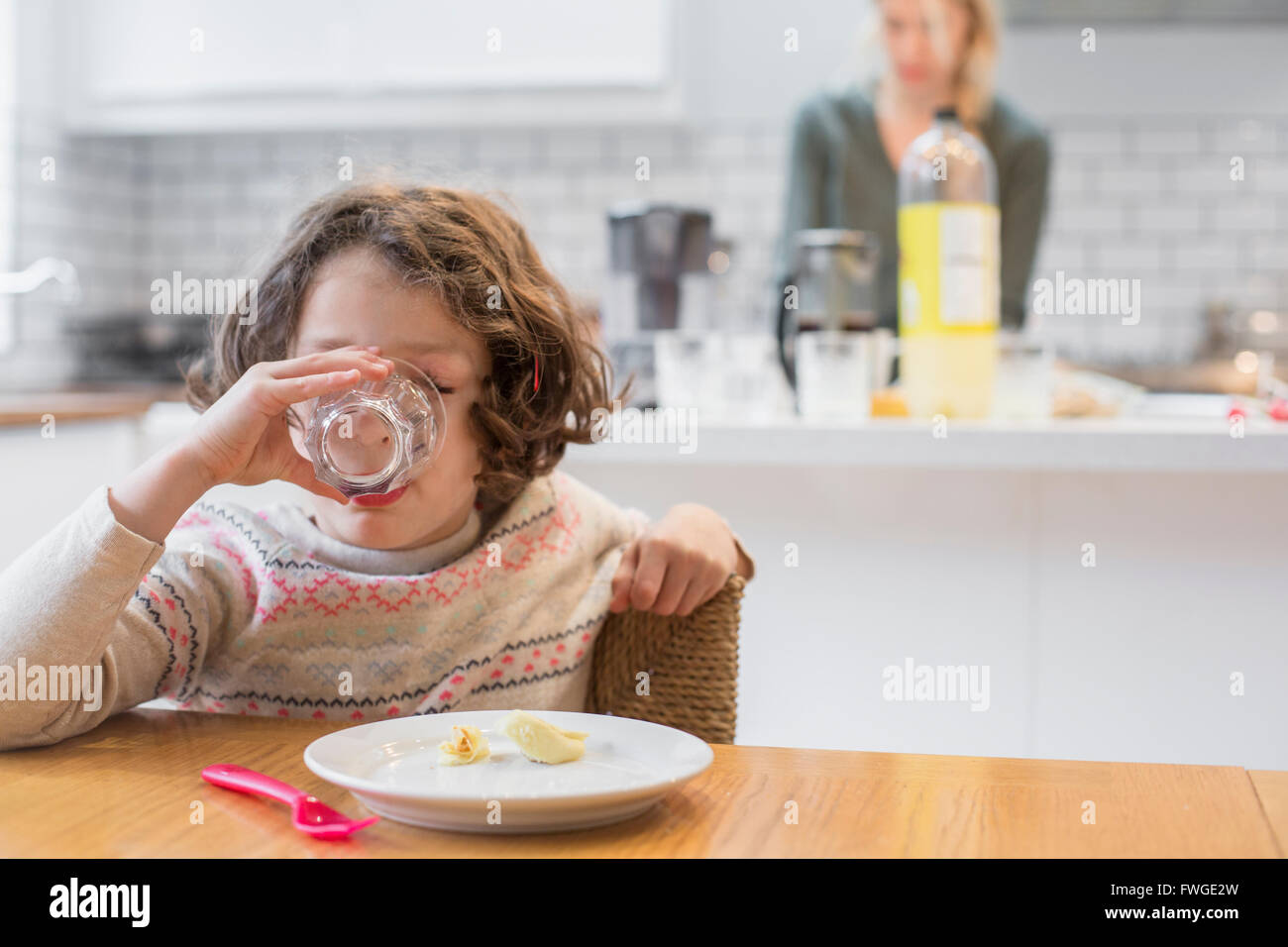 Ein Kind sitzt an einem Tisch, trinken aus einem Glas, einer Frau, hinter ihr in der heimischen Küche. Stockfoto