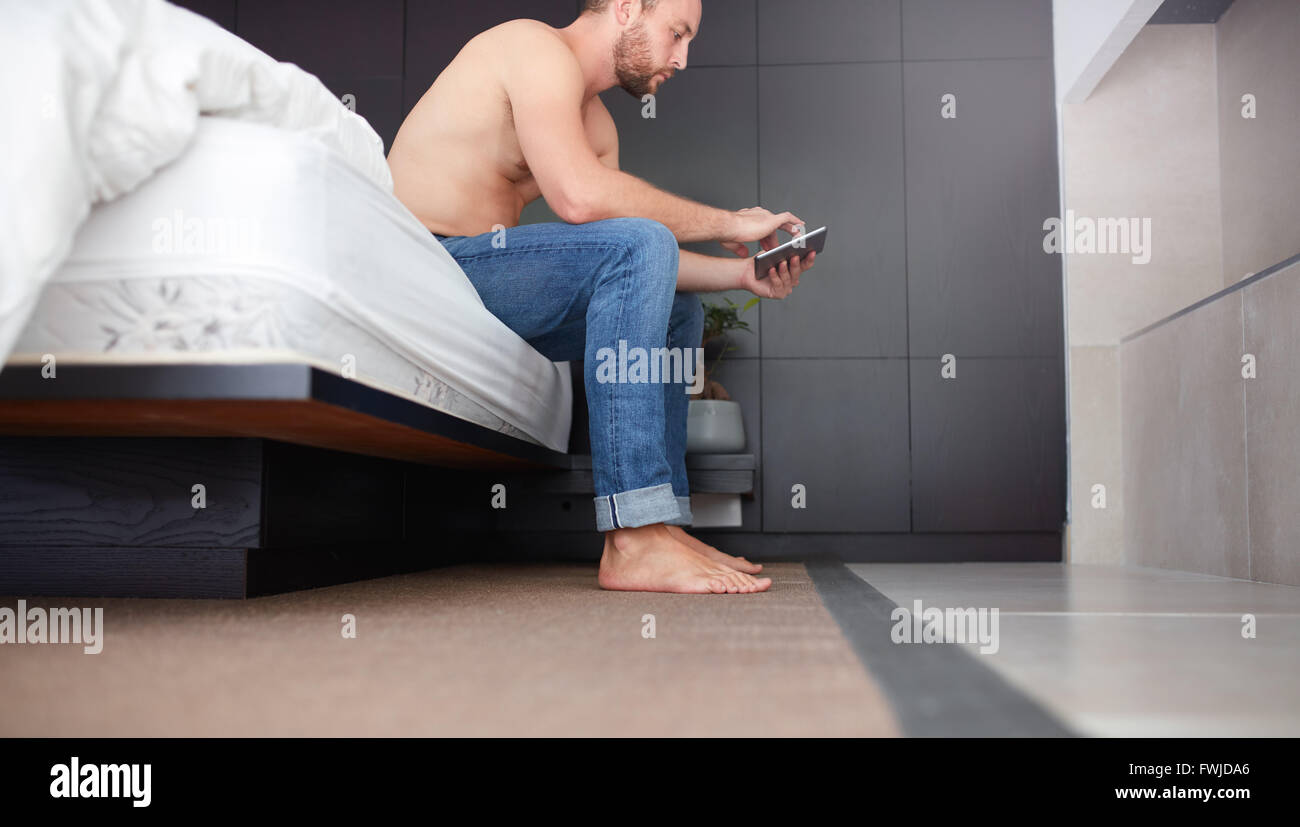 Seite Porträt eines jungen Mannes sitzt auf dem Bett im Schlafzimmer und mit digital-Tablette. Stockfoto