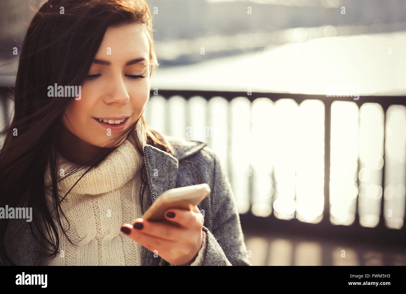 Attraktive junge Brünette Mädchen mit langen braunen Haaren und freundlichen Lächeln mit einem modernen Smartphone im Freien am hellen Frühlingstag. Stockfoto