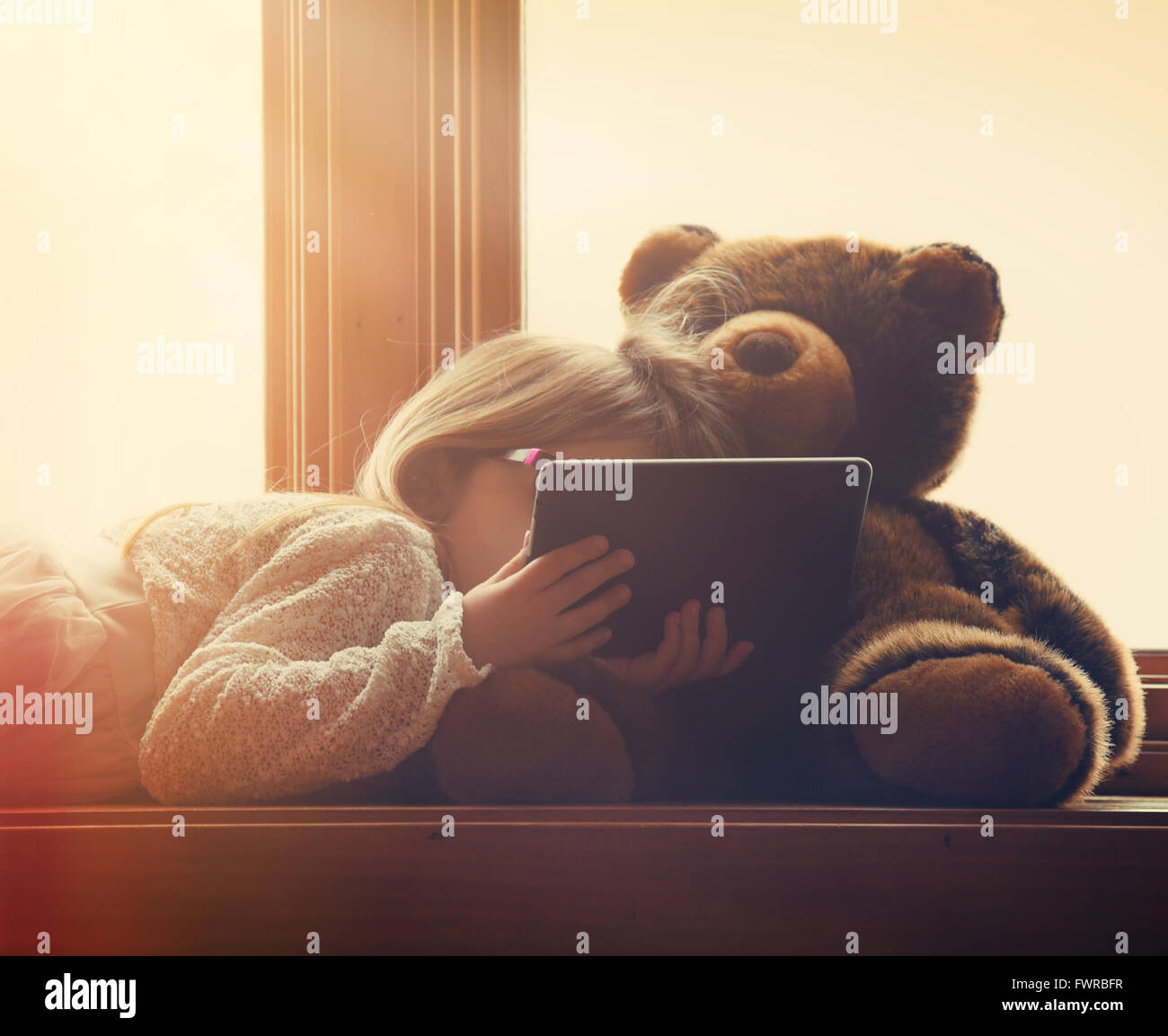 Ein kleines Mädchen hält einen Technologie-Tablet in einem sonnigen Fenster zu Hause mit einem Teddy-Bären für ein Entertainment-Freizeit-Konzept Stockfoto