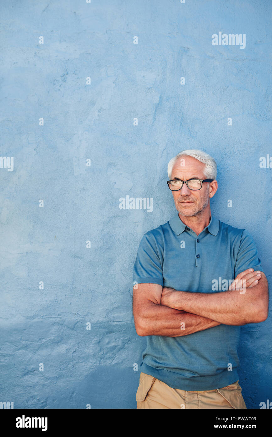 Porträt von einem Reifen kaukasischen Mann mit Brille stehend mit seine Arme verschränkt vor blauem Hintergrund. Mitte erwachsenen Mannes auf der Suche nach aw Stockfoto