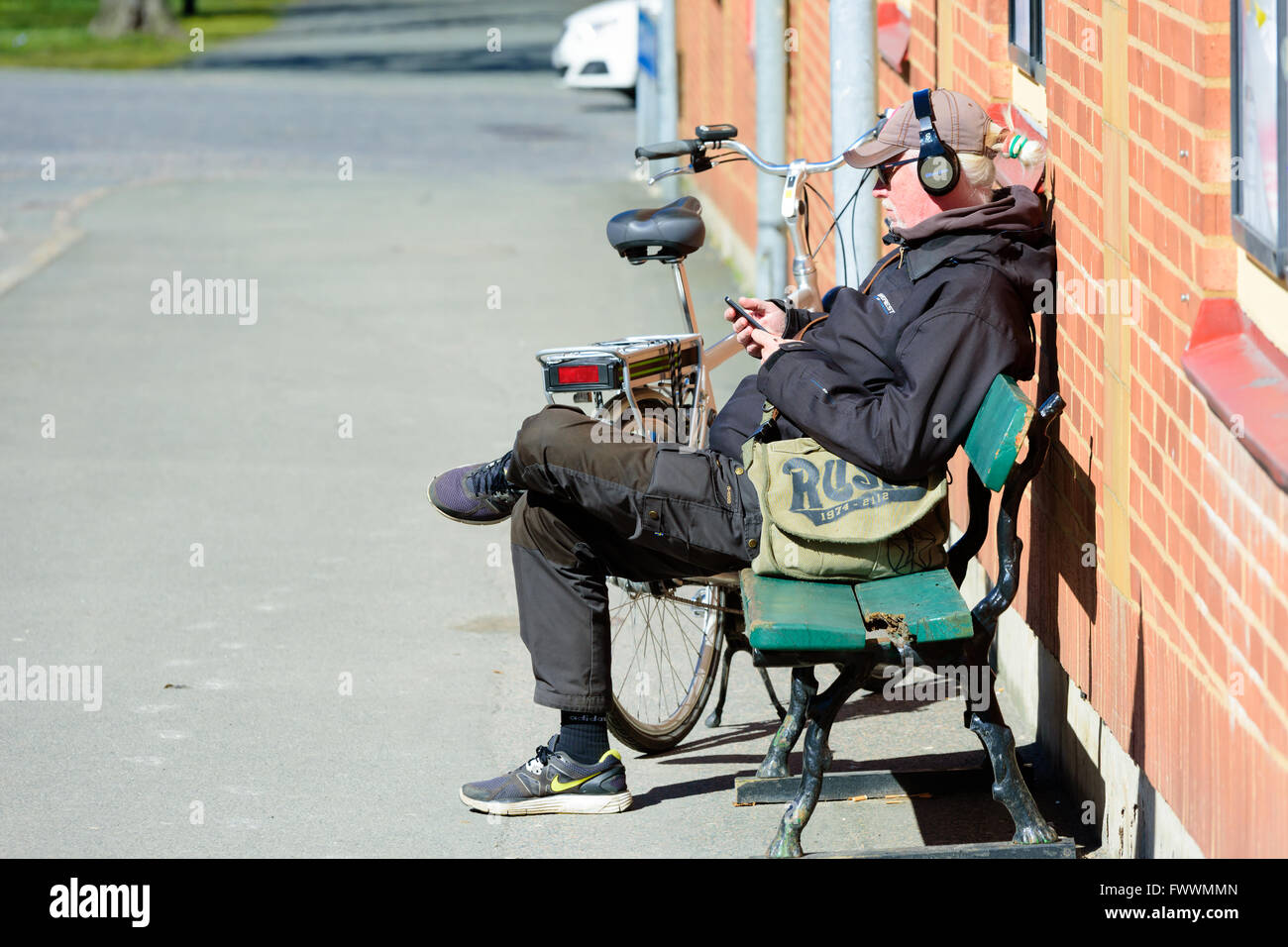 Simrishamn, Schweden - 1. April 2016: Mann sitzt auf einer Bank neben einer Wand etwas über Kopfhörer anhören. Bike ist park Stockfoto
