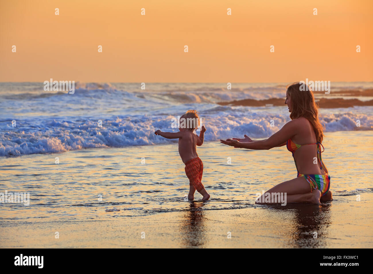 Familie Badespaß im Meer Strand Surf glückliche Mutter, Baby Sohn erster Schritt - Kleinkind laufen zu Ozeanwelle auf Sonnenuntergang Himmelshintergrund. Stockfoto