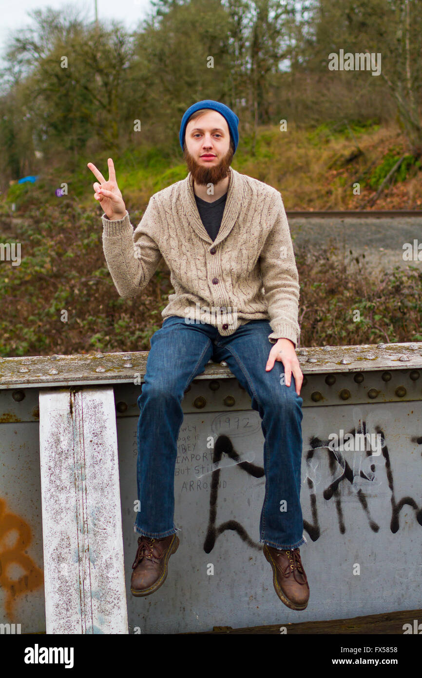 Hippie-Mann sitzt auf einer Brücke ein Friedenszeichen in diesem trendigen Hipster Fashion Porträt zu geben. Stockfoto