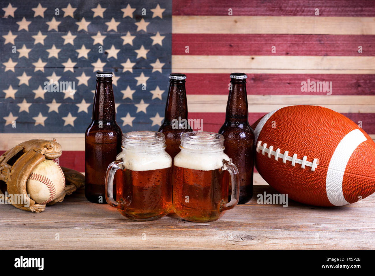 Zwei Bier Gläser gefüllt mit vollen Flaschen, Bier, Fußball und Baseball Handschuh mit Vintage Holz USA-Flagge im Hintergrund. Stockfoto