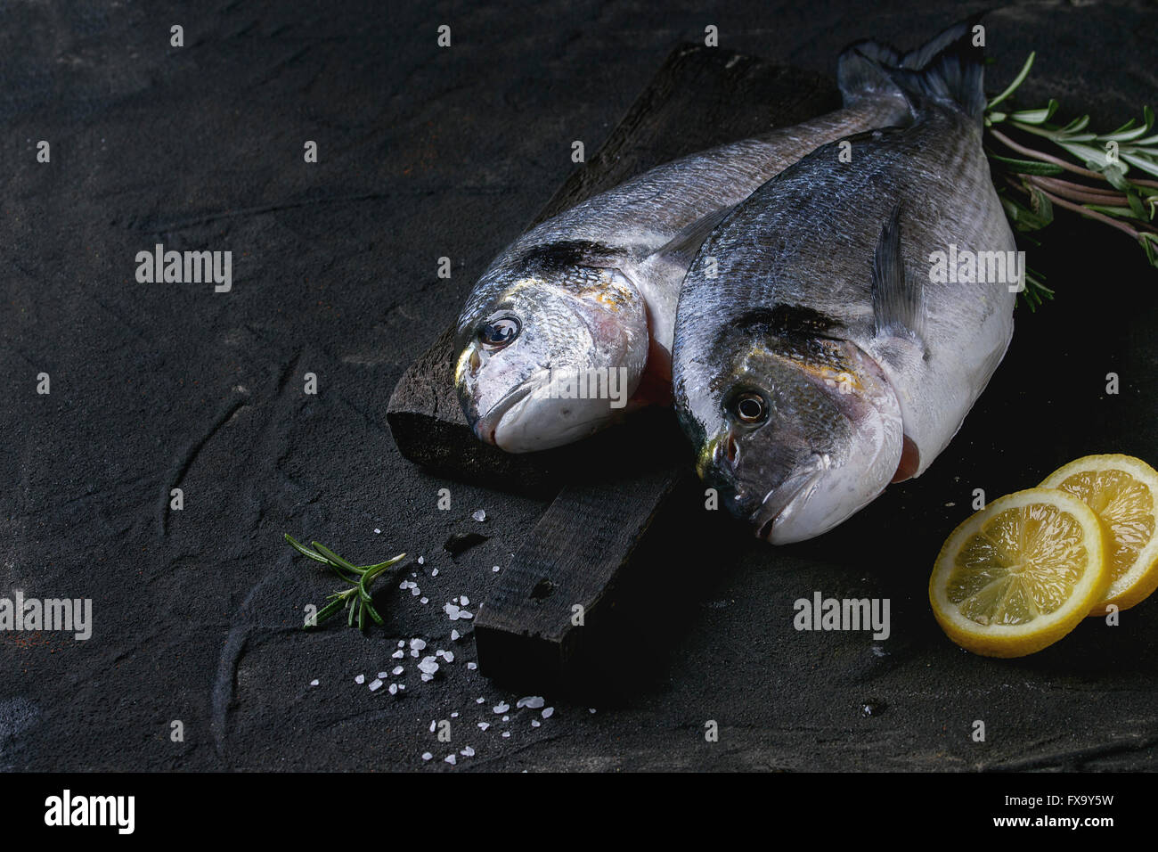 Zwei fertige rohe Brassen Fisch mit Kräutern und Zitrone auf Holzbrett über schwarz Kochen strukturierten Hintergrund. Stockfoto