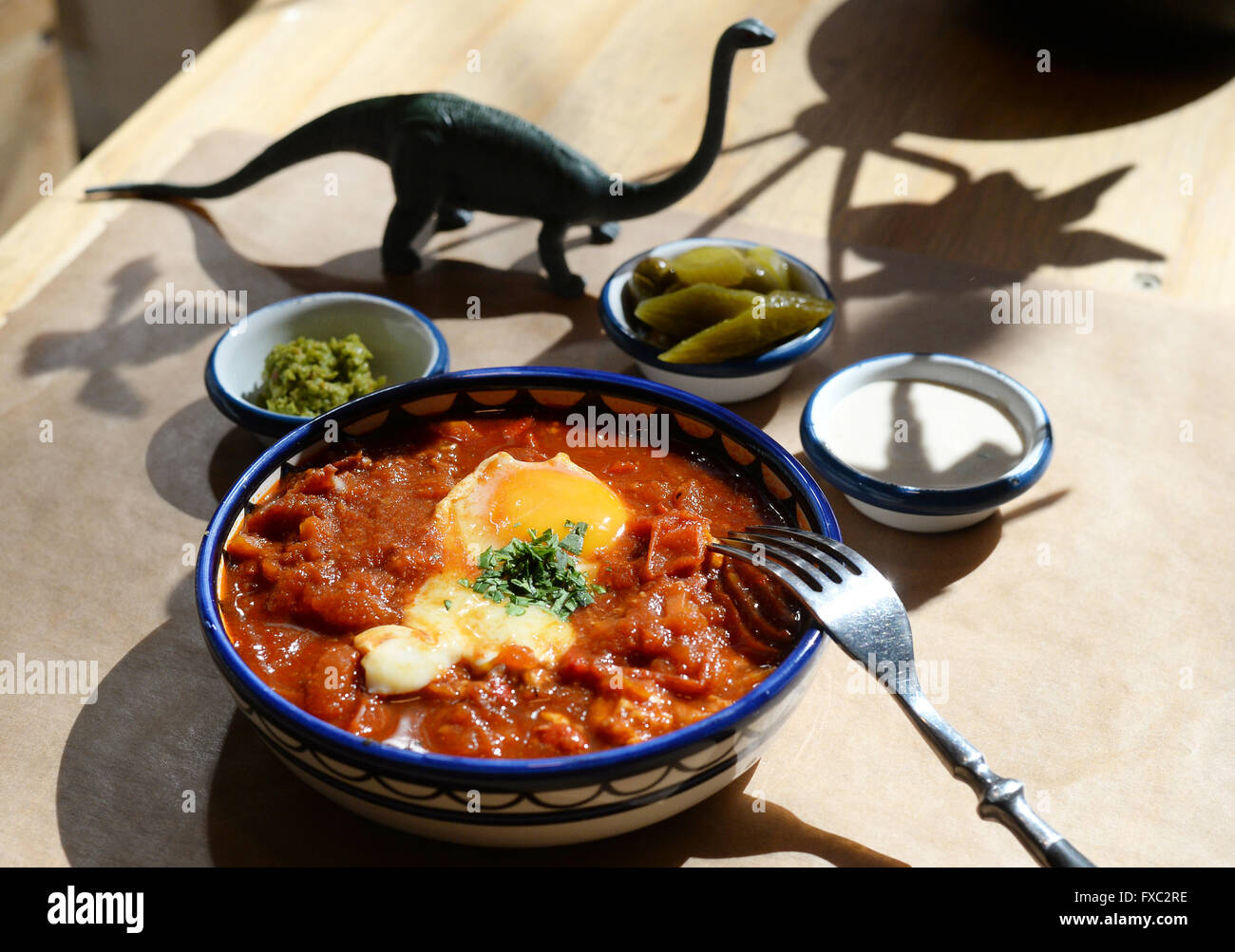 Berlin, Deutschland. 1. April 2016. Shakshuka, eine traditionelle israelische Eierspeise, sitzt auf einem Tisch mit Seiten im Cafe Gordon im Bezirk Neukölln in Berlin, Deutschland, 1. April 2016. Das Gericht wird mit pochierten Eiern in einer Soße aus Tomaten, Paprika, Chilischoten und Zwiebeln gemacht. Neben israelischen Gerichten verkaufen die Besitzer des Café, ursprünglich aus Tel Aviv, auch Aufzeichnungen aus ihrem Label Legothek. Der Dinosaurier ist der Store-Label. Foto: Jens Kalaene/Dpa/Alamy Live News Stockfoto