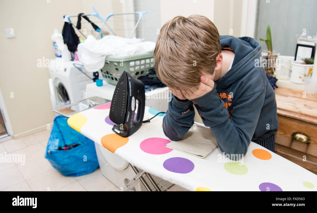 Weiße Teenager mit seinem Kopf in seinen Händen stützte sich auf eine fleckige Bügeleisen /-Brett in ein Hauswirtschaftsraum mit Waschmaschine und waschen um ihn herum Stockfoto