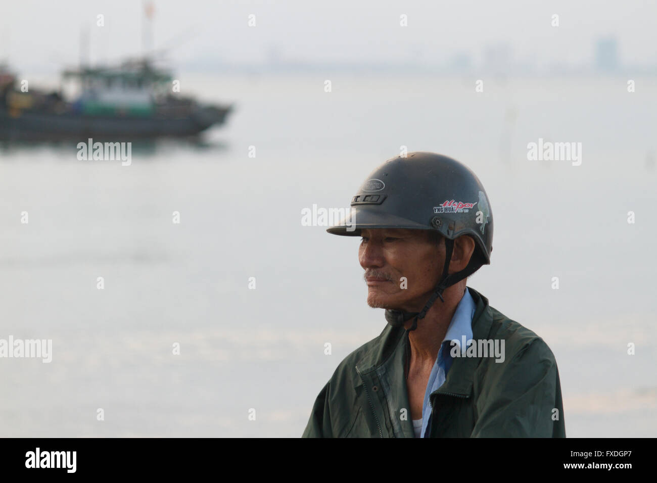 Vietnamesische Mann mit Motorradhelm vor Fischerboot und Südchinesischen Meer am Strand von Danang, Vietnam Stockfoto