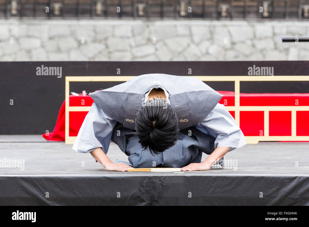 Japan, Kumamoto. Japanischer Mann kniet auf der Bühne tragen graue männliche Traditionelle yukata Jacke und Durchführen einer vollständigen tiefe Verbeugung mit Kopf fast auf dem Boden. Stockfoto