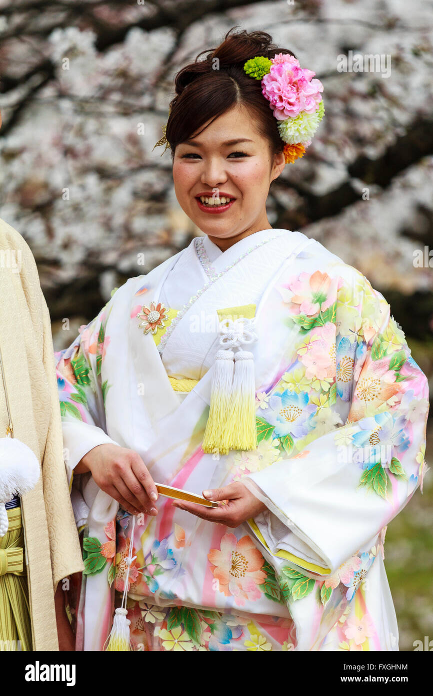 Lächelnden jungen Frau, Japanisch braut, im Kimono und Holding geschlossen Ventilator, stehend, mit unscharfen Kirschblüte im Hintergrund posiert. Frühling. Stockfoto