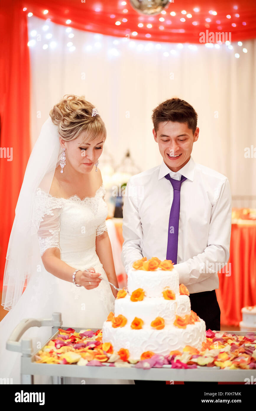 Braut und Bräutigam schneiden Sie den Kuchen bei einem Bankett mit orangen Blüten in der Dekoration. Stockfoto