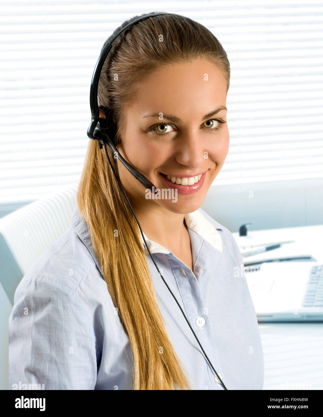 Einzelne glücklich schöne Frau mit langen Pferdeschwanz mit Telefon Kopfhörer im Call-Center oder im kleinen Büro mit Jalousien im Hintergrund Stockfoto