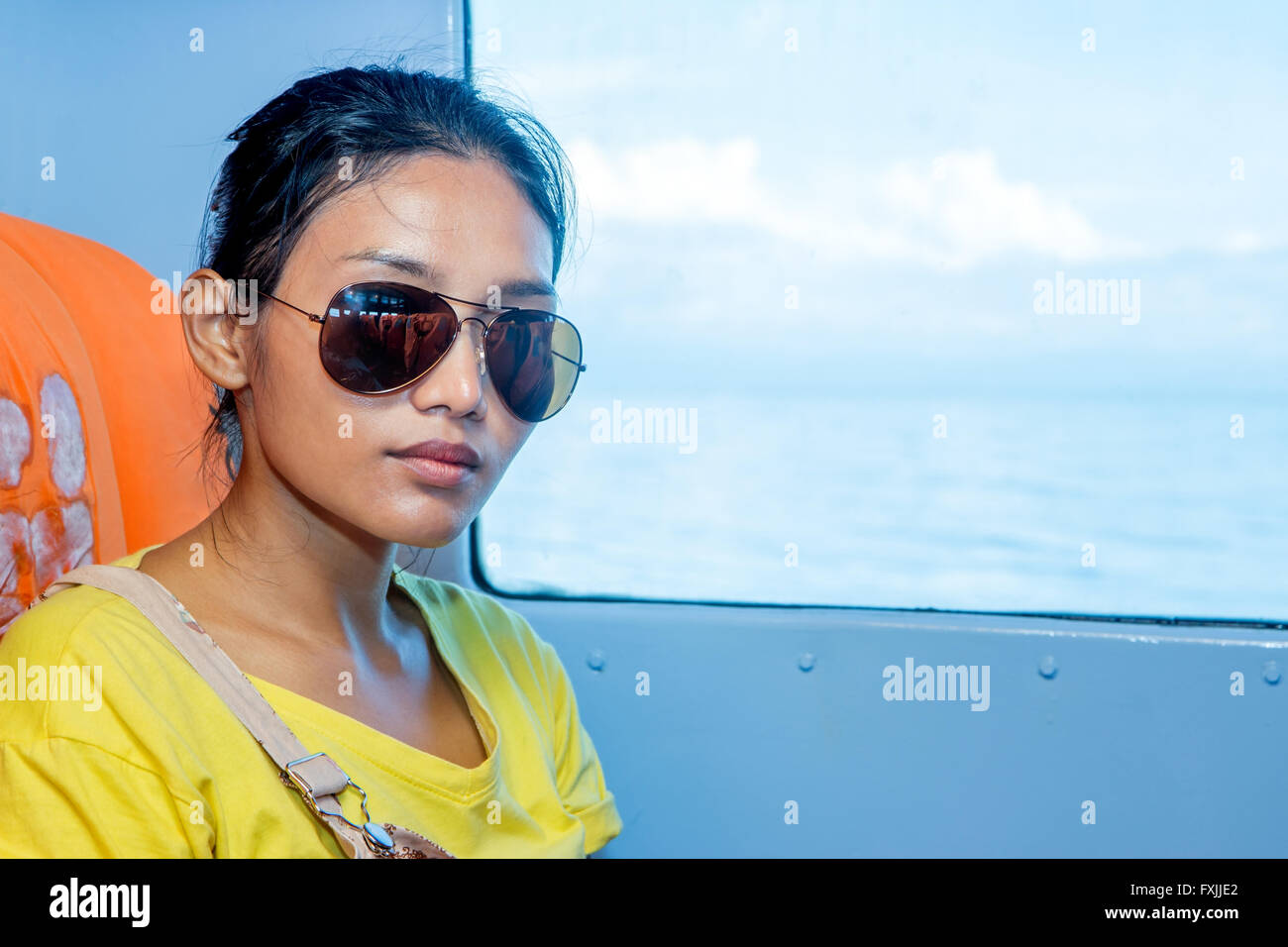 Frau mit Sonnenbrille sitzt auf einem Schiff neben Fenster mit Blick auf das Meer Stockfoto