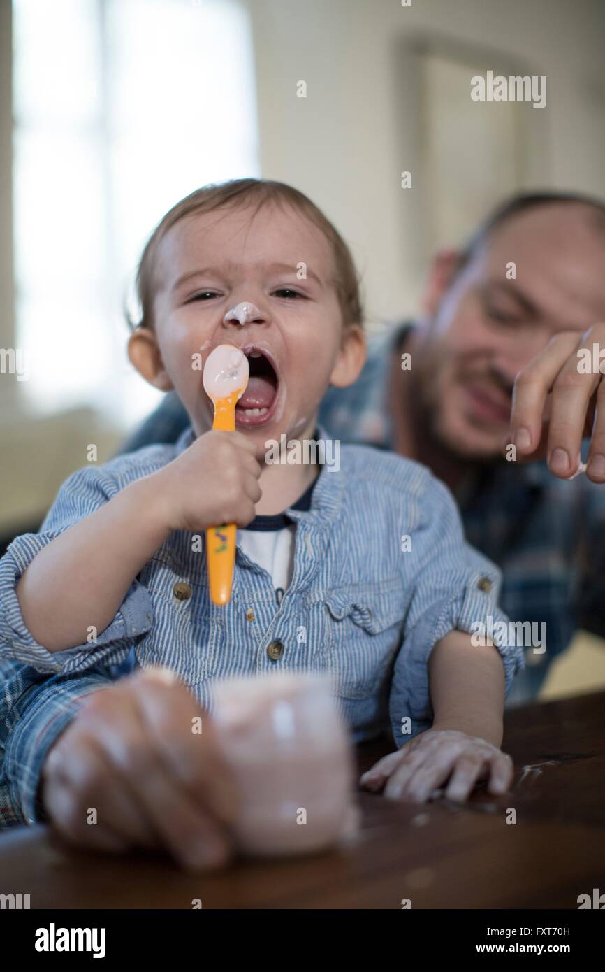 Baby Boy Fütterung selbst Joghurt mit Blick in die Kamera, offenem Mund Löffel Stockfoto