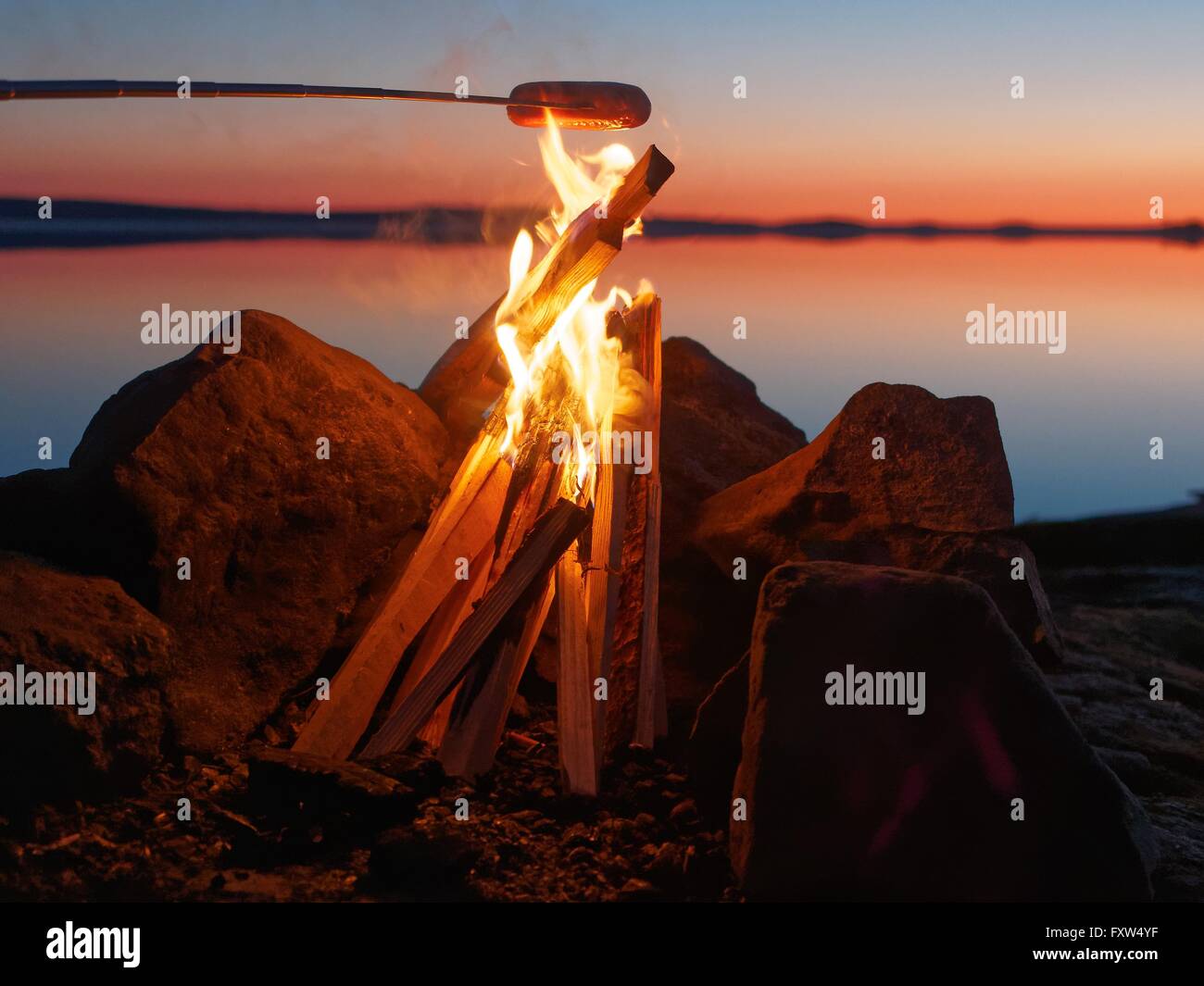 Wurst vom Grill am Lagerfeuer bei Sonnenuntergang. Stilles Wasser des Sees auf dem Hintergrund mit stimmungsvollen Abendlicht. Stockfoto