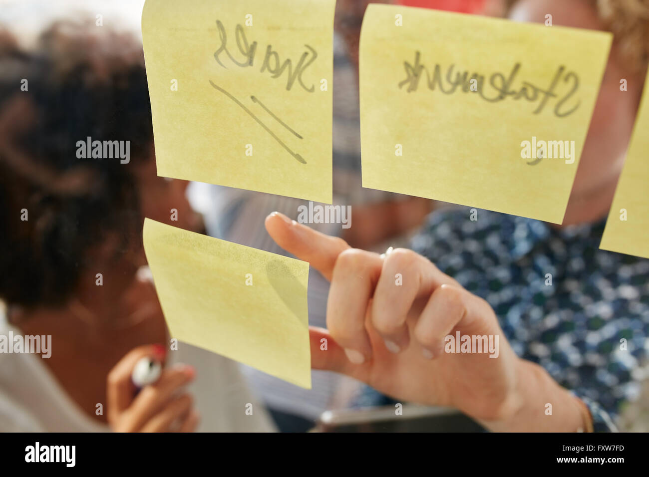 Hand der Geschäftsfrau zeigt auf eine Kurznotiz auf eine Glaswand mit Kollegen im Büro aufhalten. Fokus auf weibliche Finger. Stockfoto