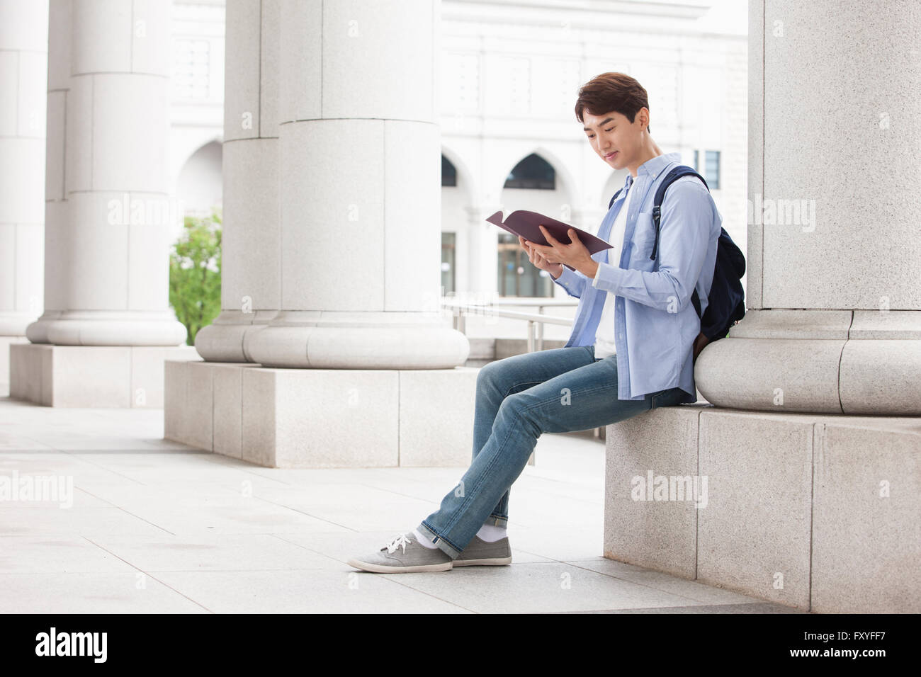 Junge College sitzt auf einer Säule auf dem Campus und liest ein Buch Stockfoto
